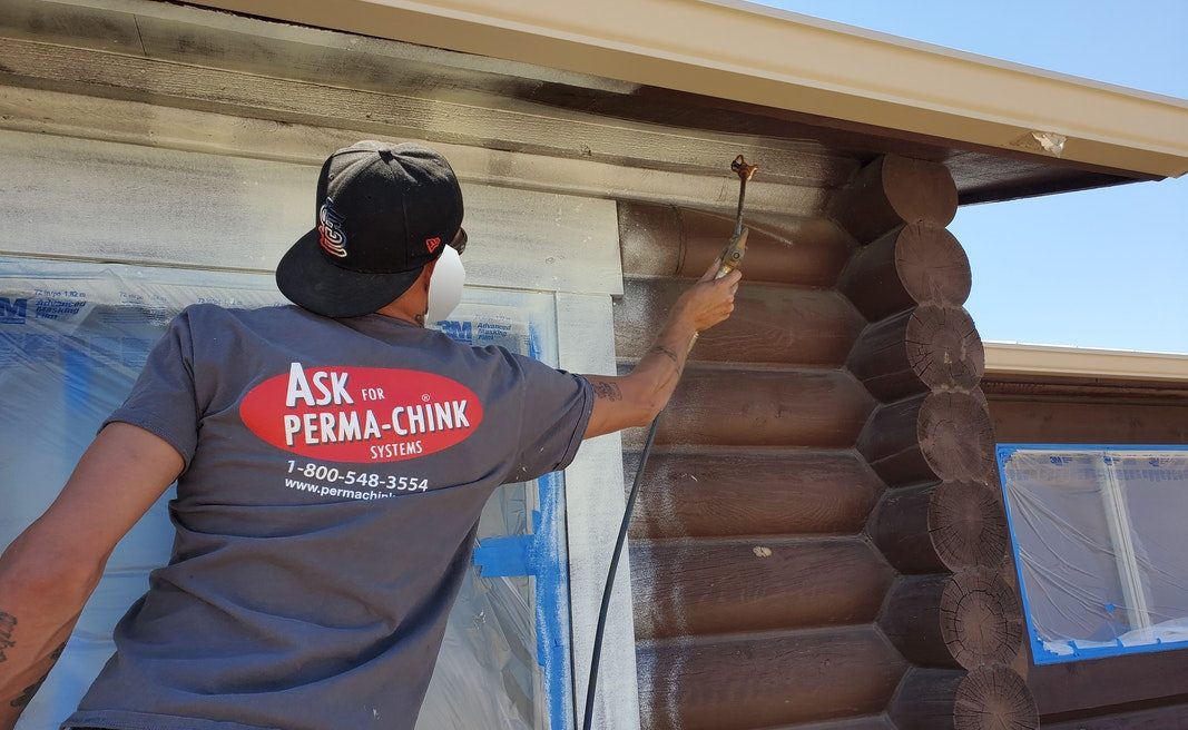 Log Master employee wearing an "Ask Perma-Chink" systems logo shirt, protective face mask, and glasses removing an old stain on a log home.